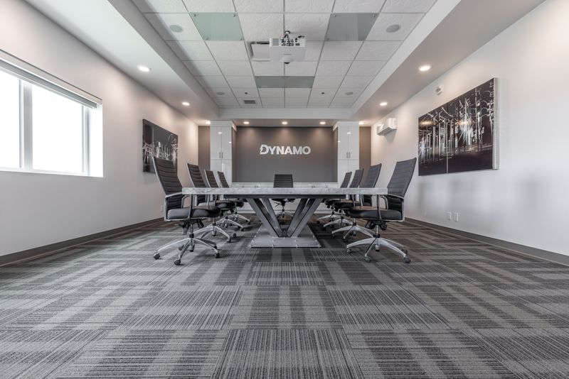 Custom-built boardroom table in the Dynamo boardroom located in Saskatoon, Saskatchewan. Dynamo logo on the back wall and 4 black chairs line each side of the table.
